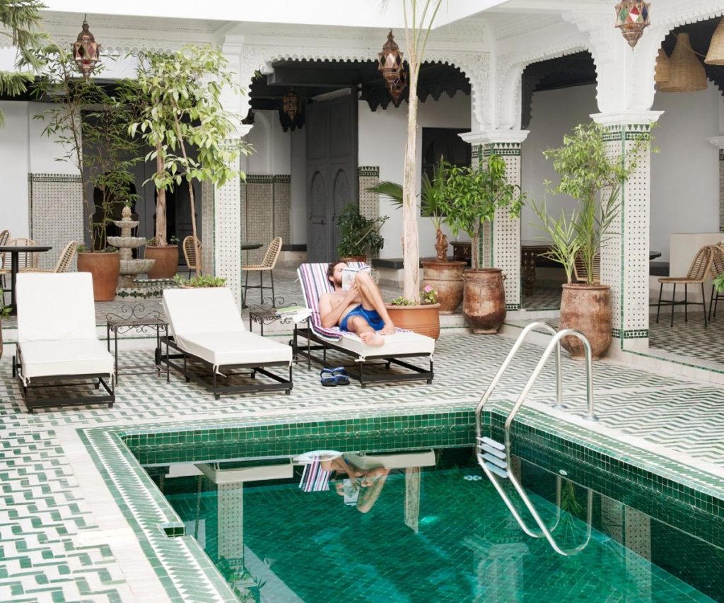 A person relaxing on a lounge chair by a swimming pool in a Moroccan-style courtyard, surrounded by potted plants and traditional architecture.