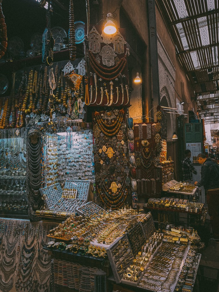 A vibrant display of jewelry and decorative items in a Moroccan souk, featuring colorful beads, intricate metalwork, and hanging ornaments, illuminated by warm light.