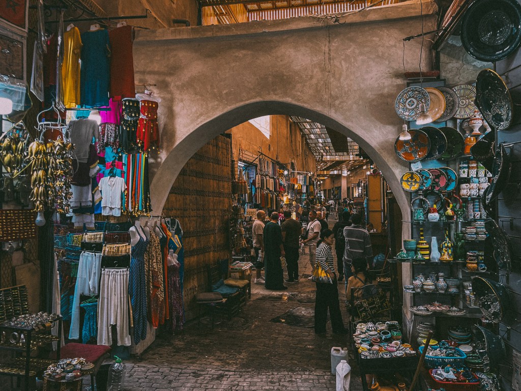 A bustling souk in Marrakesh, Morocco, featuring colorful textiles, pottery, and various artisan goods, with shoppers exploring the market.