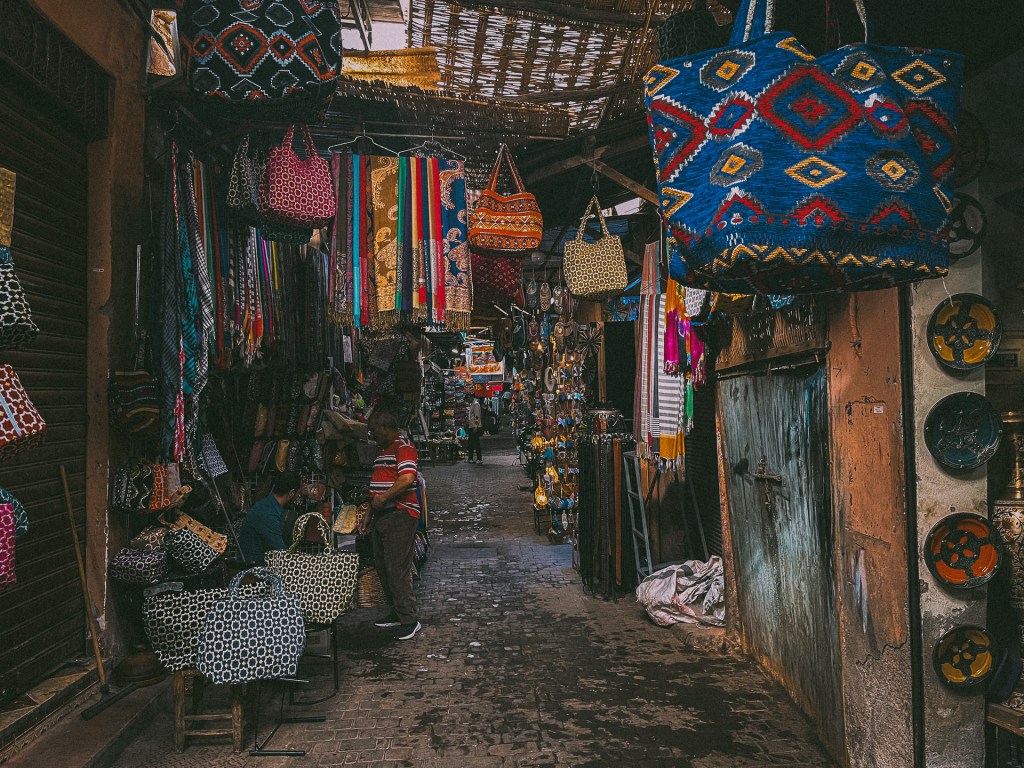 A bustling market street in Marrakesh, featuring colorful bags and textiles hanging overhead, with shoppers browsing various goods.