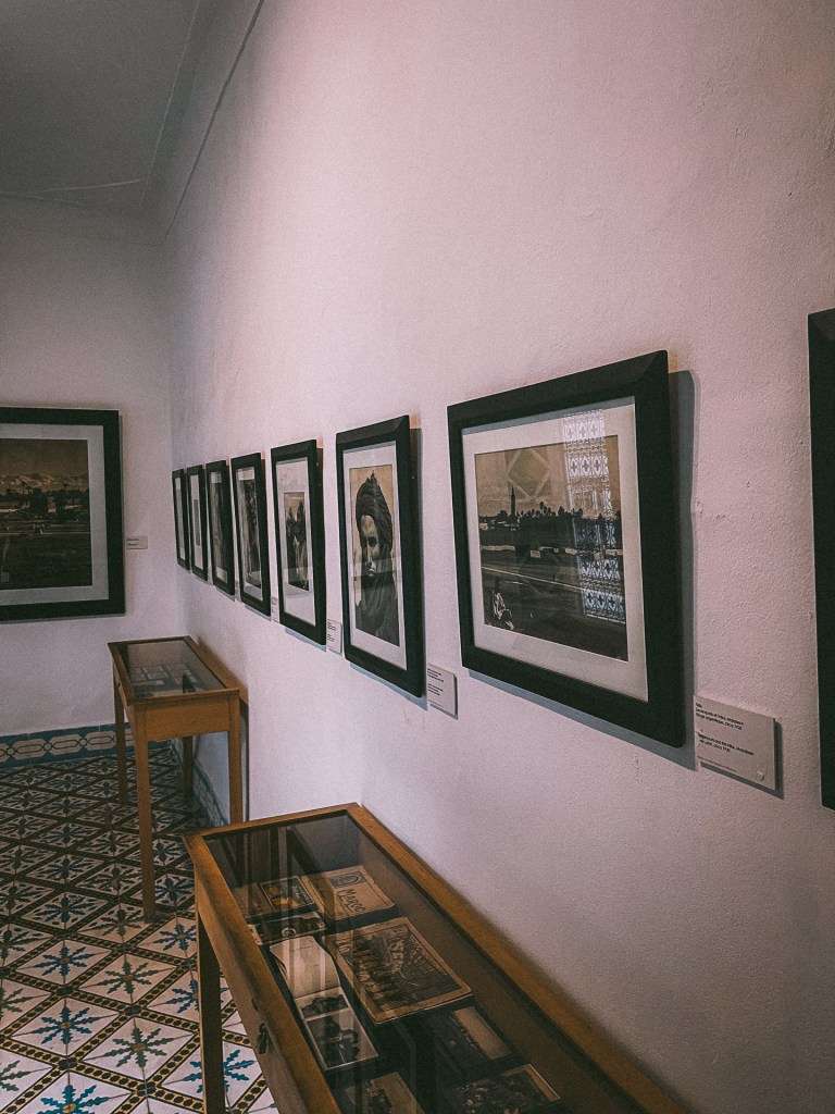 An interior view of the House of Photography in Marrakesh, featuring framed black-and-white photographs on the walls, with glass display cases containing vintage items on the floor.