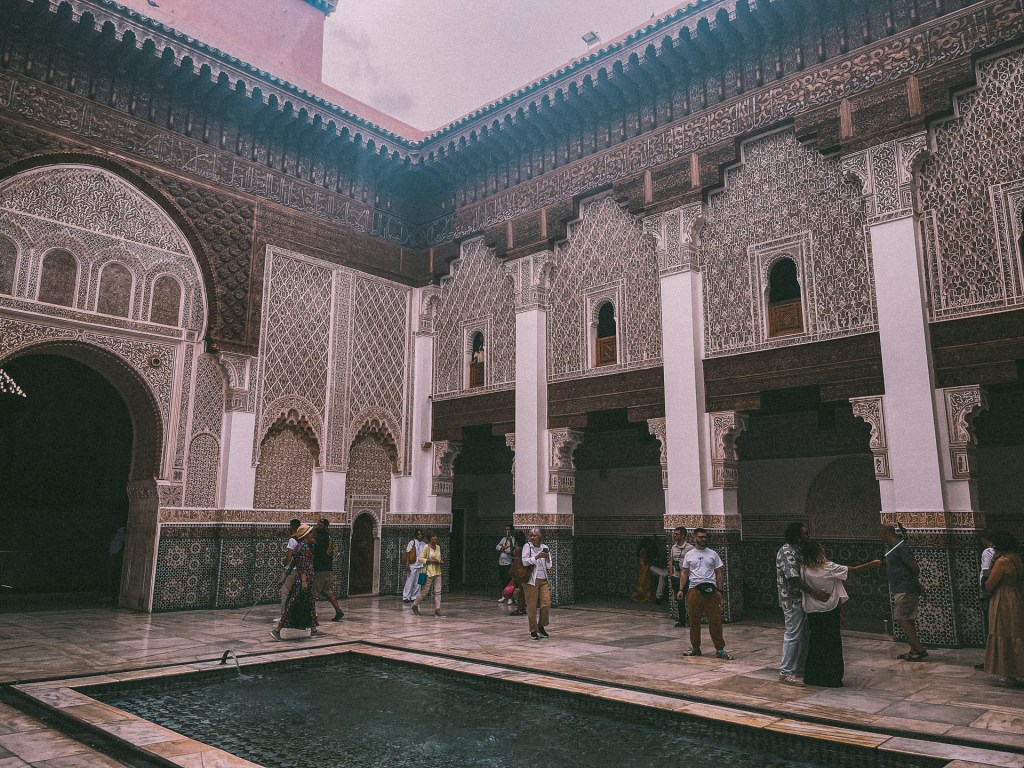 A beautifully adorned courtyard inside a Moroccan palace, showcasing intricate tilework and architecture, with visitors walking around a central pool.