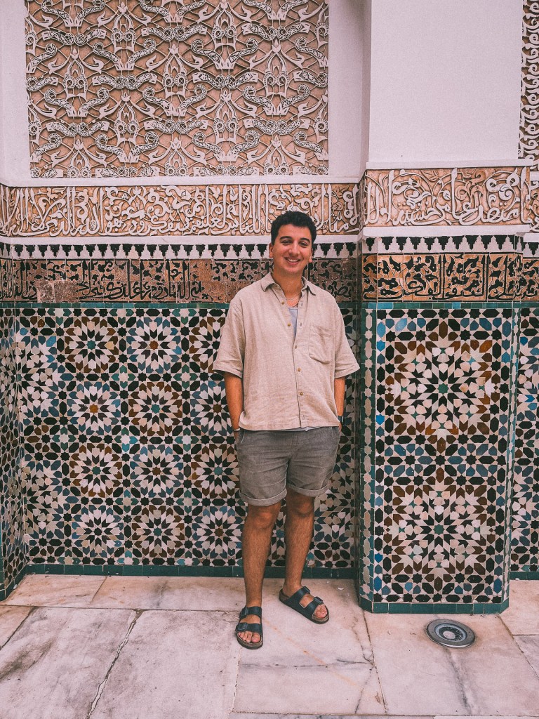 The author of this blog (me!!) standing against a beautifully decorated tiled wall with intricate geometric patterns and carved plaster designs in a Moroccan setting.