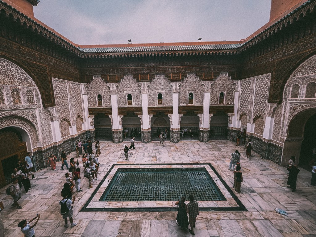A stunning view of Madrasa Ben Youssef featuring intricate tile work and arched architecture, with visitors exploring the space and a reflecting pool in the center.