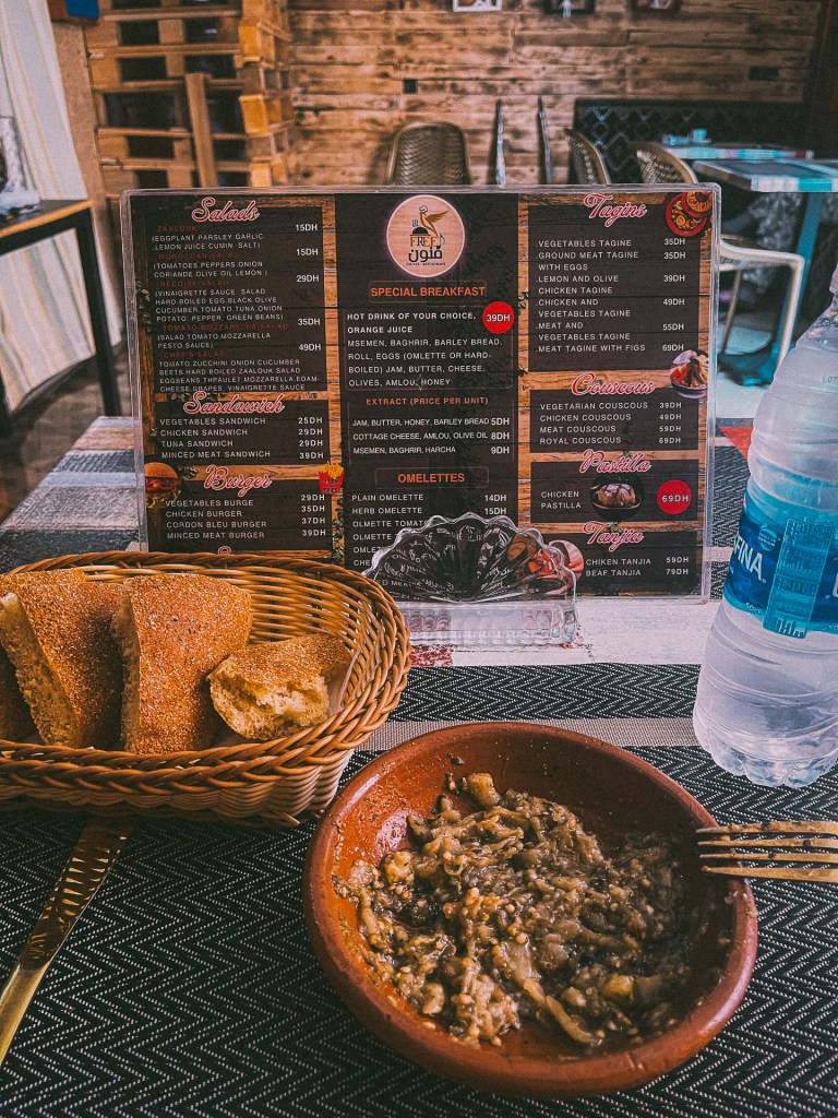 A table setting at a cafe with a menu featuring various Moroccan dishes, including salads, sandwiches, burgers, and tagines, alongside a bowl of an eggplant salad and a basket of bread.