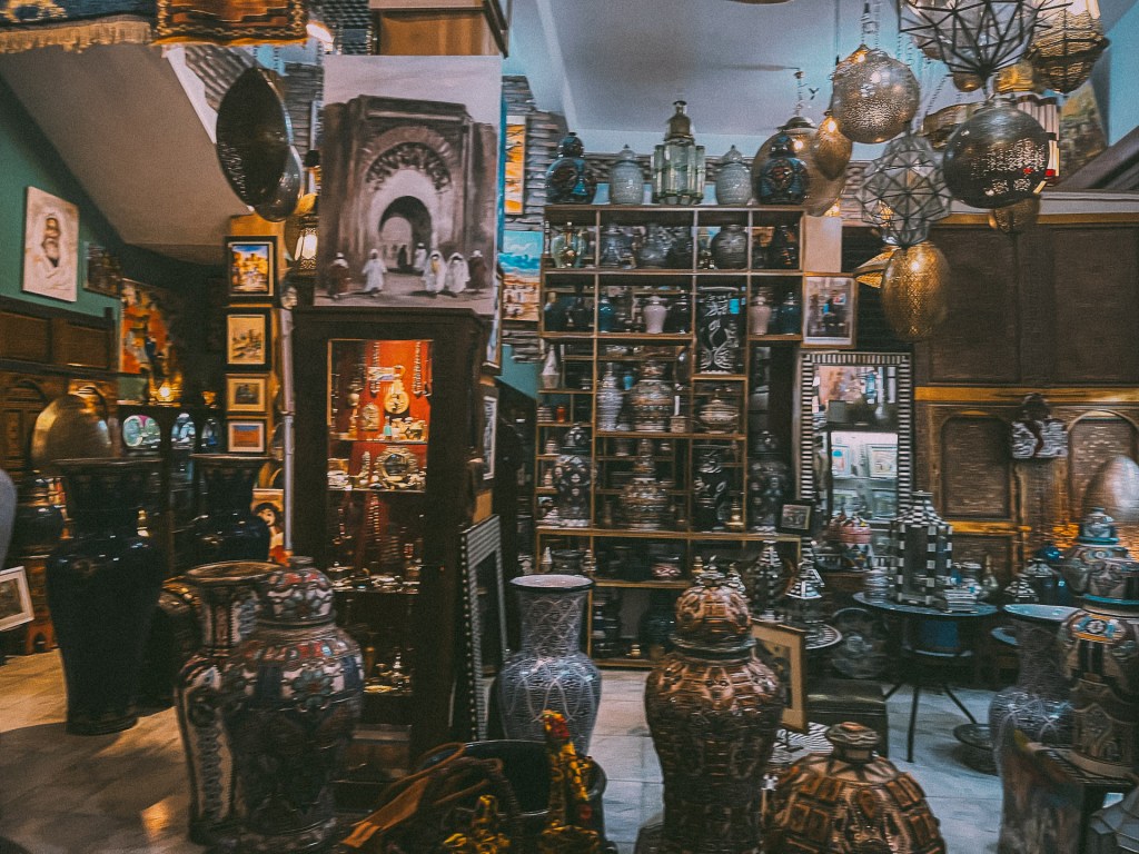An interior view of a Moroccan shop featuring intricately designed pottery, decorative lanterns, and various art pieces displayed on shelves. The walls are adorned with colorful mosaics and artworks.