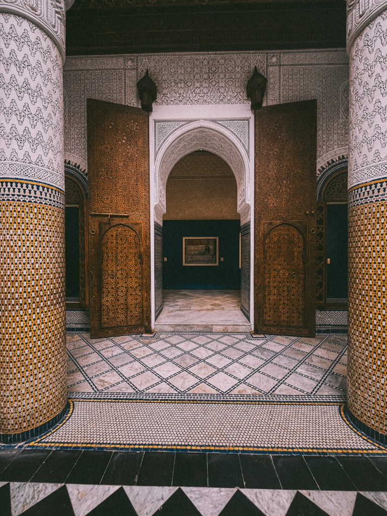 Interior view of a Moroccan architectural space featuring ornate wooden doors and intricately designed tile work on the floor and columns at Dar El Bacha Museum.