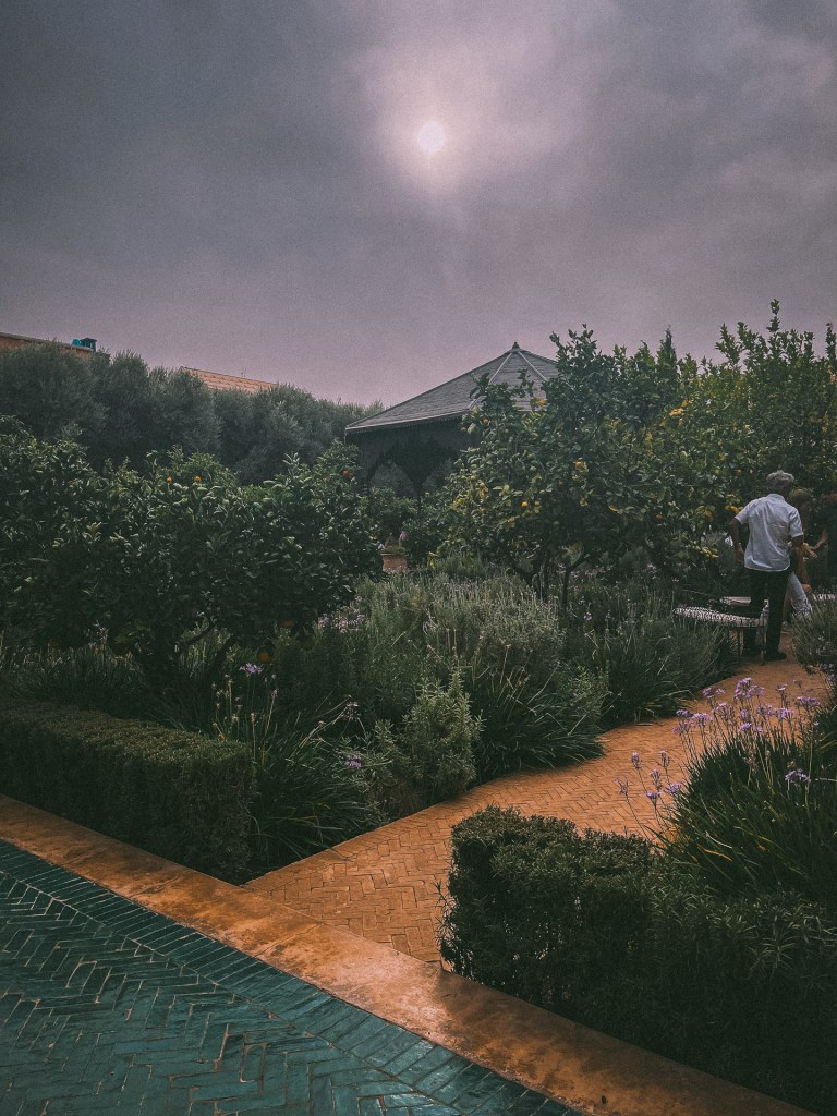A tranquil garden scene in Marrakesh featuring lush greenery, flowers, and a person walking near a gazebo under a cloudy sky at Le Jardin Secret.