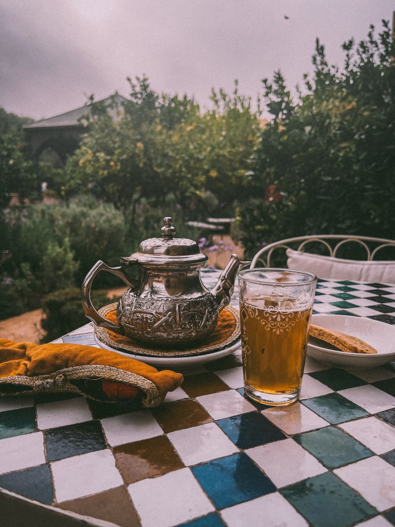 A silver Moroccan teapot sits on a colorful tiled table, accompanied by a glass of mint tea and a traditional pastry, with a lush garden backdrop at Le Jardin Secret.