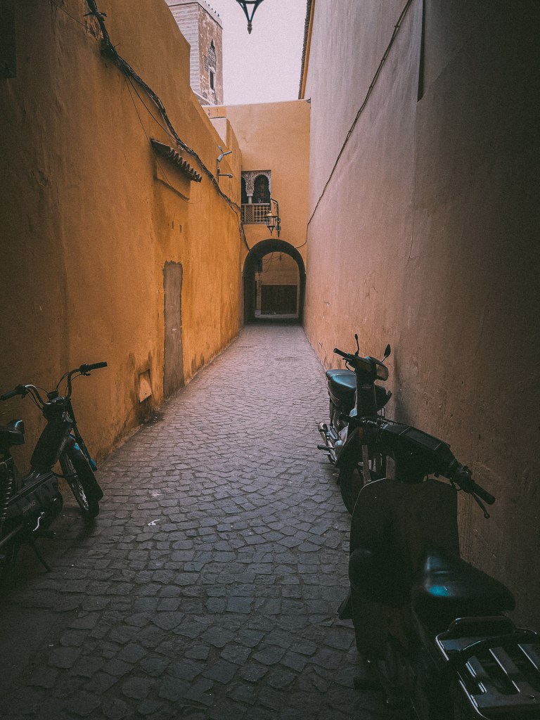 Narrow cobblestone alleyway in Marrakesh, flanked by ochre walls and parked motorcycles, leading to the Museum of Music Mouassine.