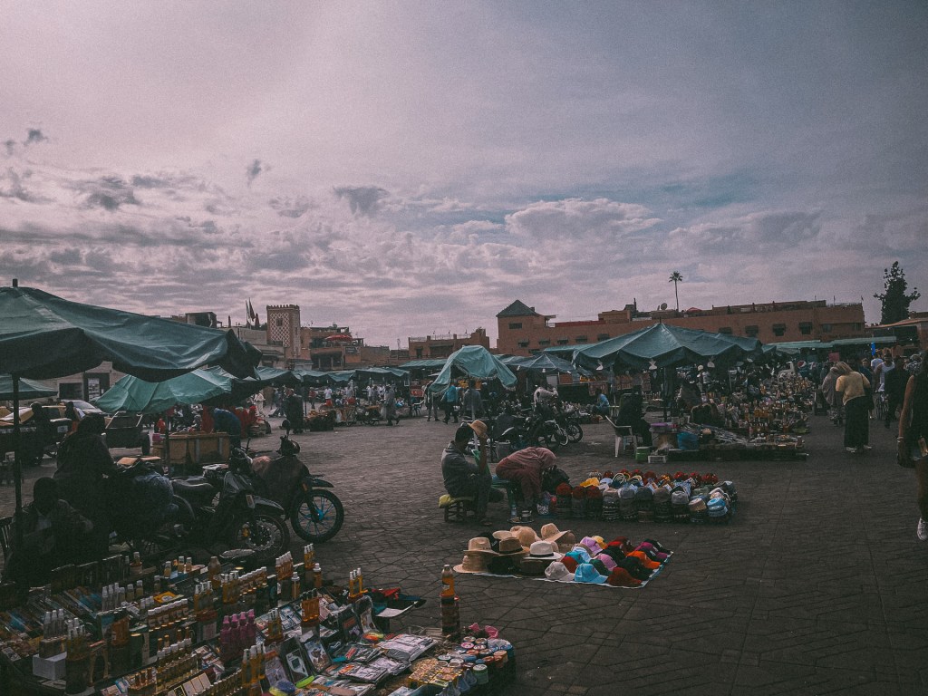 A bustling market scene in Marrakesh, specifically at Jemaa El-Fnaa, showcasing numerous stalls with colorful goods and tent canopies, under a cloudy sky.
