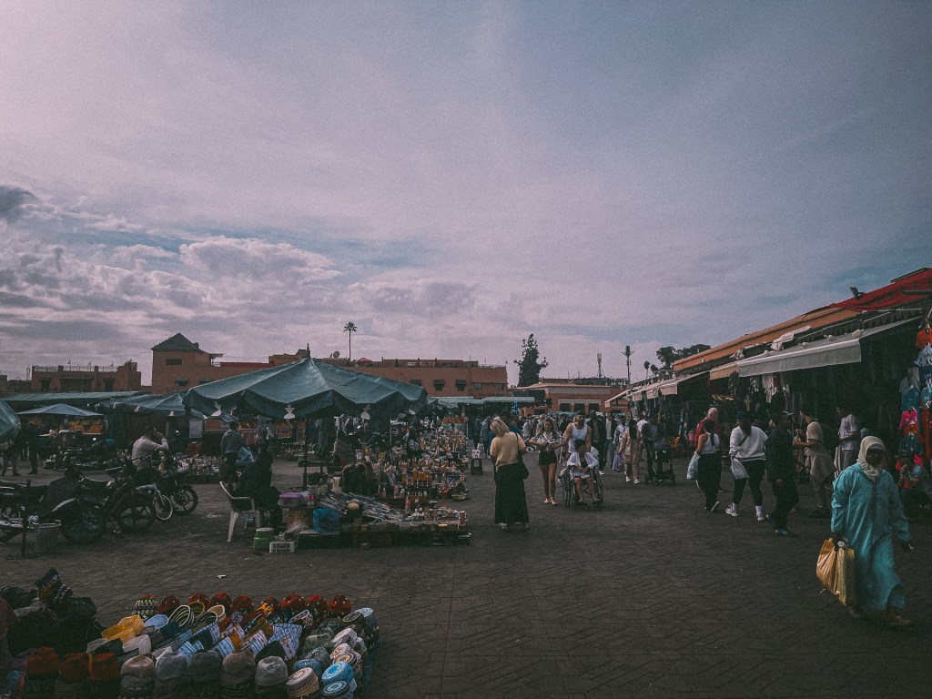 A bustling market scene in Marrakesh, with colorful stalls covered by blue canopies, displaying various goods. People are shopping and walking through the vibrant market, showcasing the lively atmosphere of Jemaa El-Fnaa.