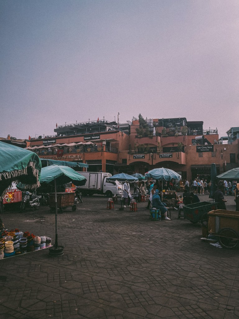 A view of a bustling market in Marrakesh, featuring colorful stalls with umbrellas, various goods for sale, and buildings at Jemaa El-Fnaa.