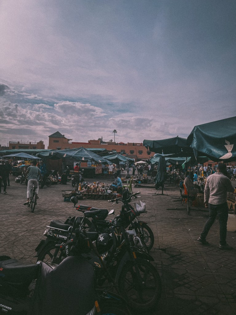 A bustling market scene in Marrakesh with people shopping and interacting among various stalls and canopies, while bicycles are parked in the foreground at Jemaa El-Fnaa.