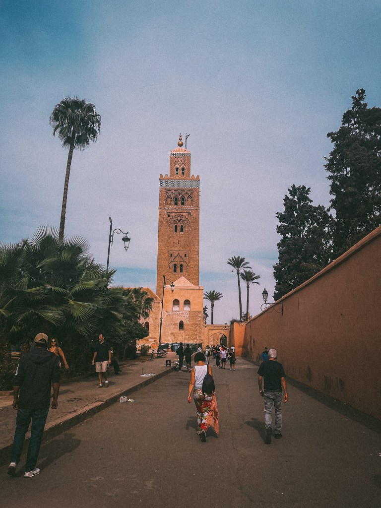 View of the Koutoubia Mosque in Marrakesh with people walking along a pathway lined with palm trees.