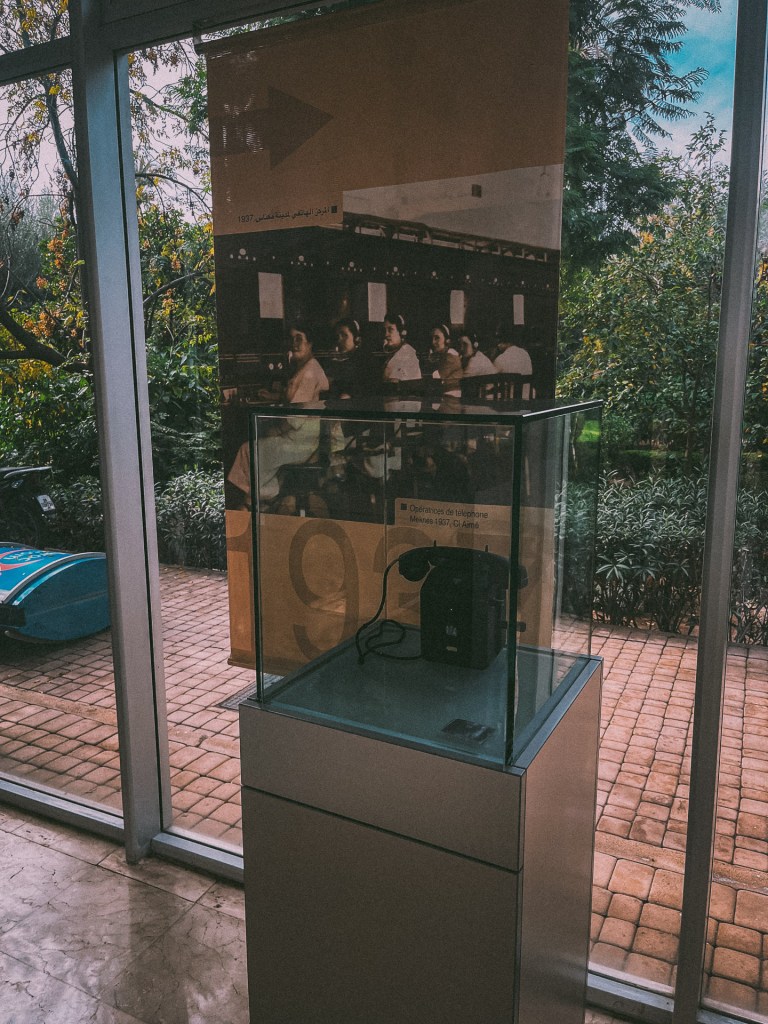 A display featuring a historical telephone model, with a background showcasing a black and white photo of women at a telecommunications station. The setting is at the Telecom Museum with green plants visible outside the glass entrance.