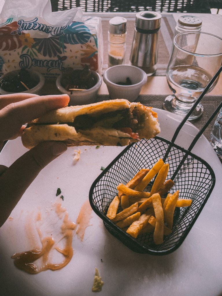 A hand holding a panini filled with various toppings, accompanied by a small metal basket of golden French fries on a table set with condiments and a glass.