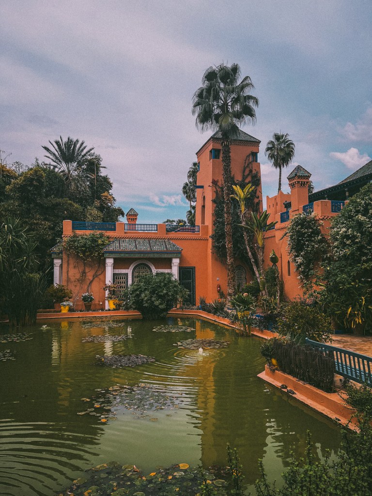 A picturesque view of the Jardin Majorelle in Marrakech, featuring vibrant orange buildings surrounded by lush greenery and a tranquil pond.