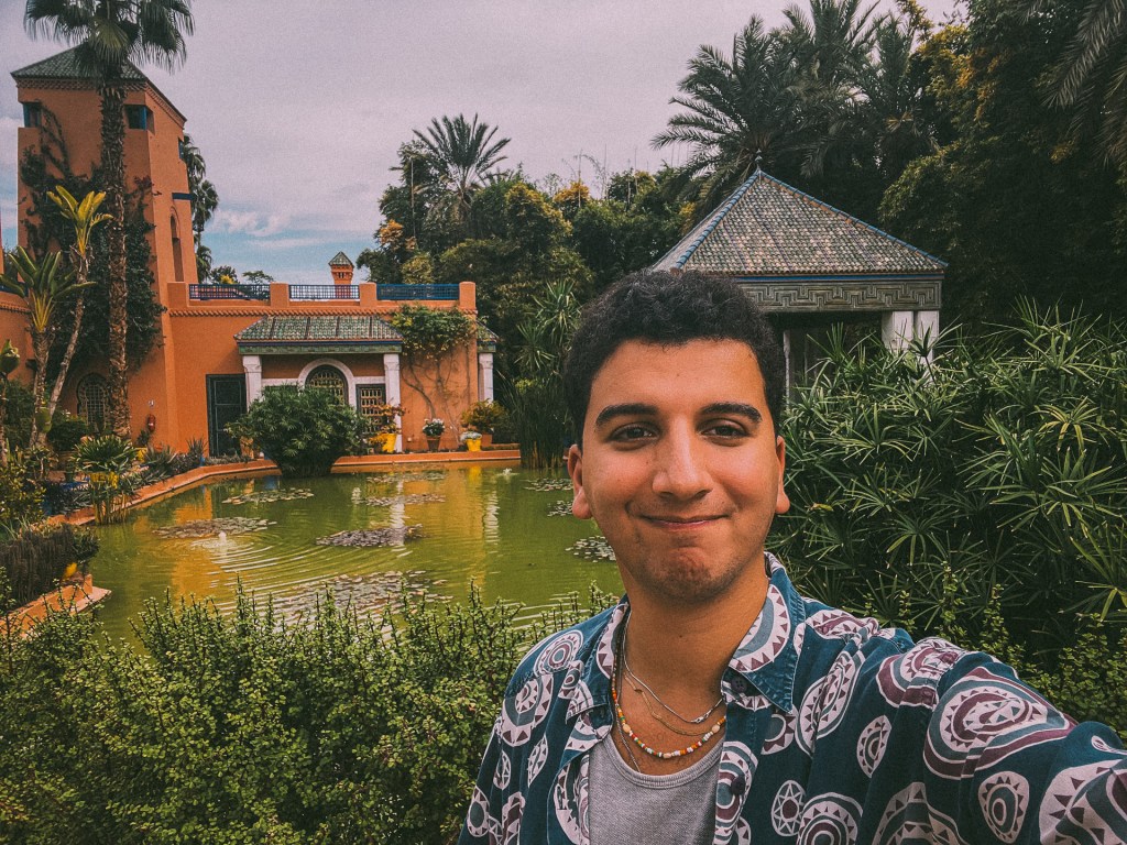 A selfie in a lush garden setting, featuring palm trees, a small pond, and an architectural structure with a tiled roof. And me!