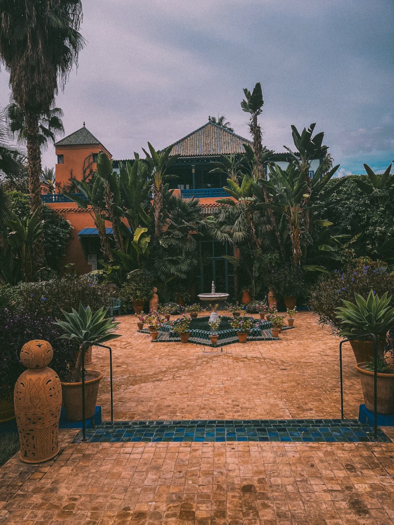 A picturesque view of Jardin Majorelle featuring palm trees, a central fountain surrounded by flower pots, and traditional architecture in the background.