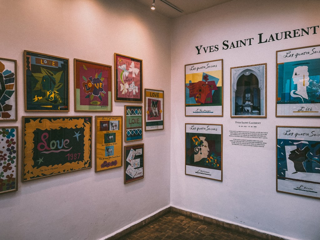 An exhibition wall featuring colorful framed artworks with the word 'Love' and seasonal themes, displaying the influence of Yves Saint Laurent, showcased at the Yves Saint Laurent Museum.
