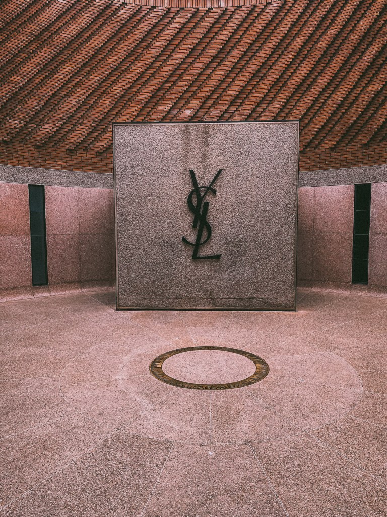 Interior view of the Yves Saint Laurent Museum in Marrakesh, featuring a black 'YSL' logo against a textured wall, surrounded by a circular stone floor and a brick ceiling.