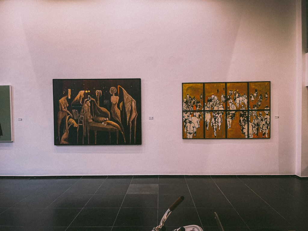 An art gallery interior showcasing two paintings. The left painting features abstract figures, while the right painting depicts a group of people in motion, both set against a minimalist wall at the Loft Art Gallery Marrakech.