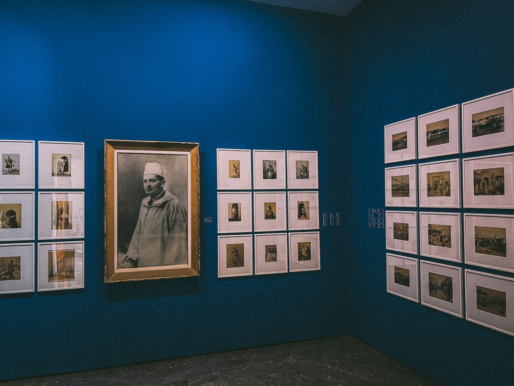 An exhibition room at the MACMA Museum featuring various framed photographs on a blue wall, including a prominent portrait of a man in traditional clothing.