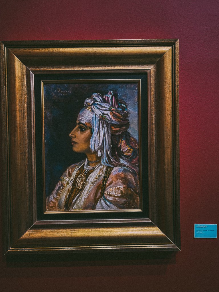 A framed painting depicting a woman in traditional Moroccan attire, showcased against a deep red wall at the MACMA Museum.