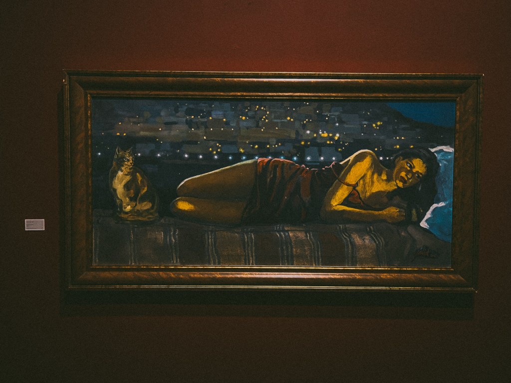 Painting of a woman at the MACMA Museum lying on a bed next to a cat, set against a dark background with glowing lights.