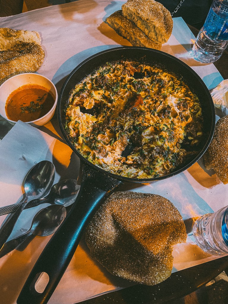 A traditional Moroccan dish served in a black skillet, surrounded by bread and a small bowl of sauce, on a tabletop with utensils and a bottle of water.
