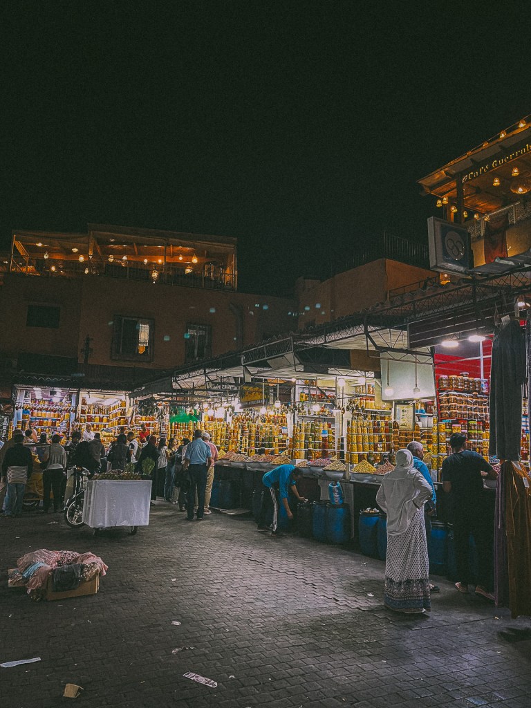 A bustling night market in Marrakesh, featuring vibrant stalls filled with jars of spices and local products, surrounded by shoppers and visitors.