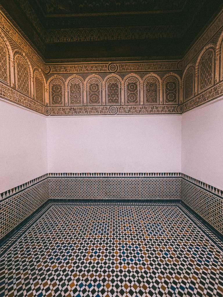 Interior view of a Moroccan room at the Bahia Palace featuring intricate geometric tile work on the floor and detailed decorative patterns on the walls and ceiling.