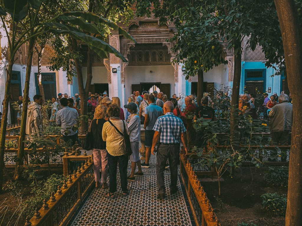 A busy courtyard at the Bahia Palace filled with tourists surrounded by lush greenery and traditional Moroccan architecture.