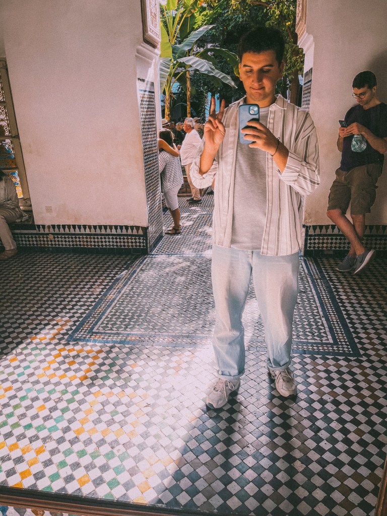 Me taking a selfie in a decorative tiled hallway, surrounded by plants and other visitors at the Bahia Palace.