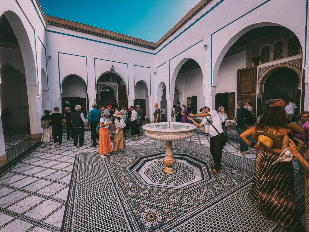A bustling interior of Bahia Palace featuring a beautiful fountain surrounded by intricate tile work, with several tourists exploring the space and engaging with each other.
